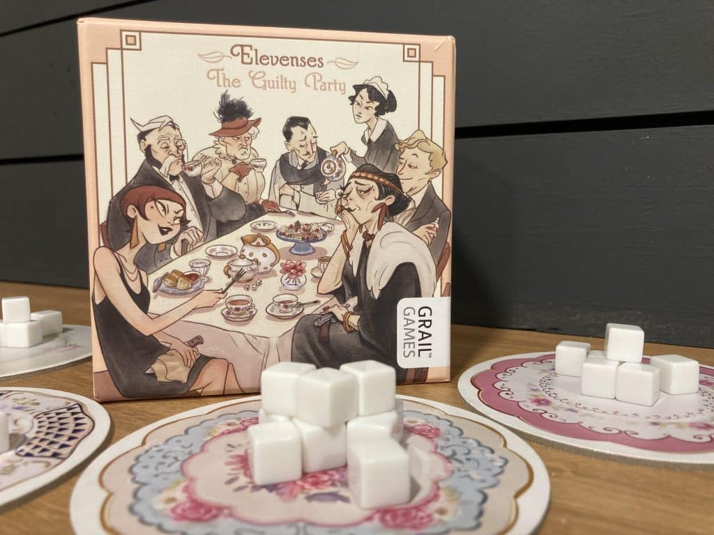 Elevenses: The Guilty Party board game box cover with plates filled with small white acryllic sugar cubes in front of it.